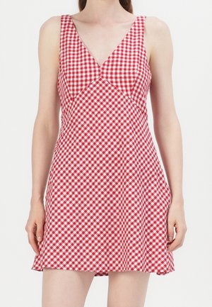 Mujer con un vestido sin mangas de cuadros rojos y blancos con escote en V y falda acampanada sobre un fondo liso.