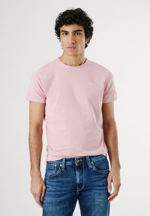 Light pink cotton t-shirt with short sleeves and a round neckline, featuring small white logo detail on the left chest. Worn with blue jeans.