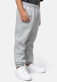 Niño con pantalones de chándal grises y zapatillas blancas, de pie con las manos relajadas a los lados frente a un fondo simple.