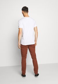 Homme debout de dos, portant une chemise blanche à manches courtes, un pantalon marron et des baskets noires basses sur un fond blanc uni.