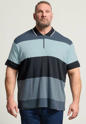 Uomo con barba che indossa una polo a maniche corte con righe orizzontali blu e nere, in piedi davanti a uno sfondo chiaro uniforme.