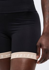 Gros plan sur une personne portant un short noir avec des revers beiges affichant le mot « ENDLESS » en lettres blanches.