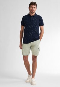 Námornícky polokošelový vrch s drobným vzorem, spárovaný se světle zelenými cargo šortkami a bílými teniskami. Polokošile má klasický límec a krátké rukávy.