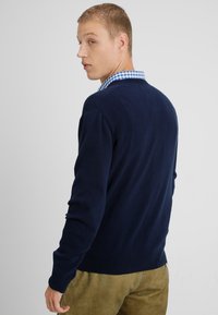 Marineblauer Pullover mit geripptem Saum und Bündchen, über einem karierten blau-weißen Hemd mit Kragen getragen. Glatte Textur, taillierte Passform.