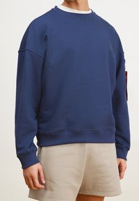 Felpa blu navy con scollo a girocollo, polsini e bordo a coste. Presenta una vestibilità comoda e un piccolo logo sul petto. Abbinata a pantaloni corti beige.