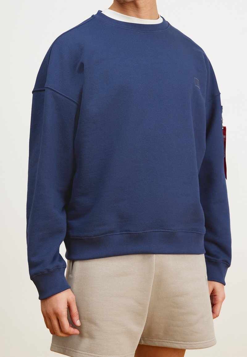 Felpa blu navy con scollo a girocollo, polsini e bordo a coste. Presenta una vestibilità comoda e un piccolo logo sul petto. Abbinata a pantaloni corti beige.