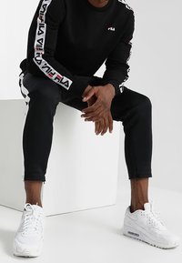 Chemise noire à manches longues avec logo FILA blanc et rouge sur les manches, associée à un pantalon noir ajusté et des baskets blanches de sport.