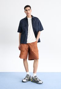 Camisa de manga corta azul marino, camiseta blanca, pantalones cortos de carga naranja, zapatillas grises con acentos amarillos. Conjunto casual sobre un fondo liso.