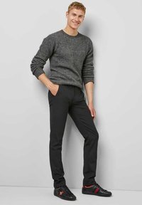 Grijze gebreide trui, zwarte strakke broek en zwarte sneakers met rode accenten. Model staat met handen in de zakken voor een neutrale achtergrond.