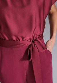 Bordowa bluzka o gładkiej, satynowej fakturze, z marszczonym pasem i wiązaną akcentacją. Krój jest luźny i swobodny.