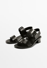 Sorte hælede sandaler med krokodillemønster, der har to brede remme og en metalspænde, på en lav blokhæle. Glat sål.