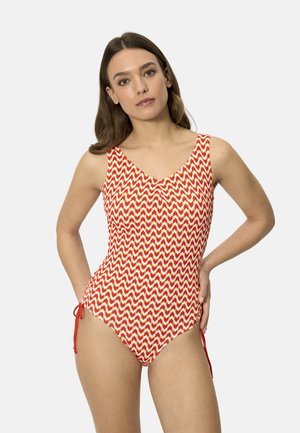 Mujer con cabello largo y castaño que lleva un traje de baño de una pieza rojo y blanco con patrón de chevrones y lazos laterales, de pie con las manos en las caderas.