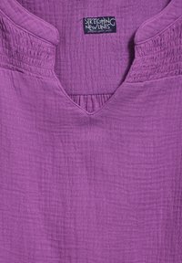 Haut violet en tissu texturé avec un décolleté en V et des accents froncés aux épaules. Présente une étiquette de marque "Stretching New Lines."