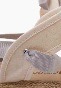 Sandales en toile beige avec des bretelles en tissu gris et une semelle en jute tissée. Les lanières se nouent à la cheville et présentent un petit détail en métal en boucle.