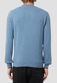 Uomo che indossa un maglione a maniche lunghe azzurro chiaro e pantaloni neri, visto di spalle su uno sfondo grigio chiaro uniforme.