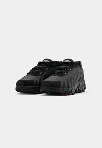 Sapatos desportivos pretos com um upper de malha texturizada, sola grossa com recortes redondos e área de atacadores reforçada com detalhes reflexivos.