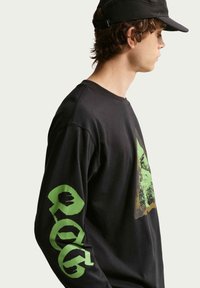 Νεαρός άνδρας με μαύρο καπέλο και μαύρο φούτερ με πράσινο γραφικό στο στήθος και μεγάλο πράσινο κείμενο στο μανίκι, εμφανίζεται σε προφίλ σε ανοιχτό φόντο.