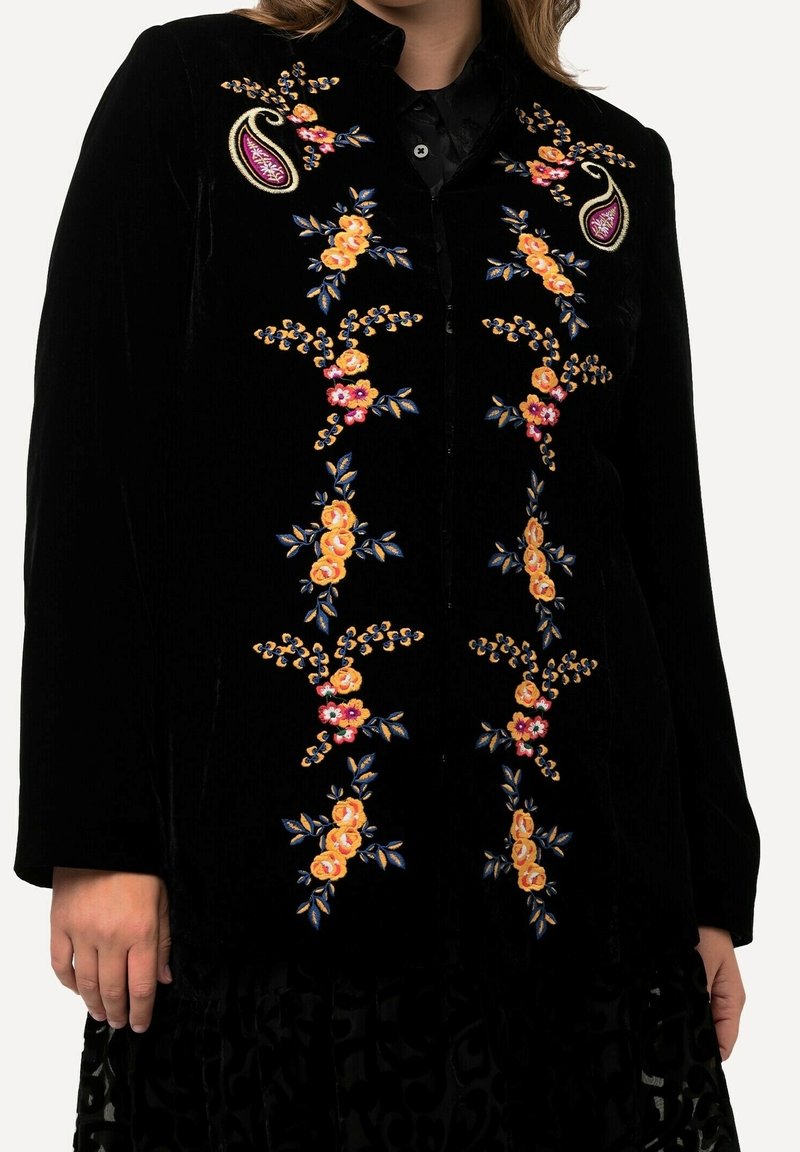 Veste en velours noir avec broderie florale verticale symétrique en jaune, rose et vert, portée sur une chemise noire.