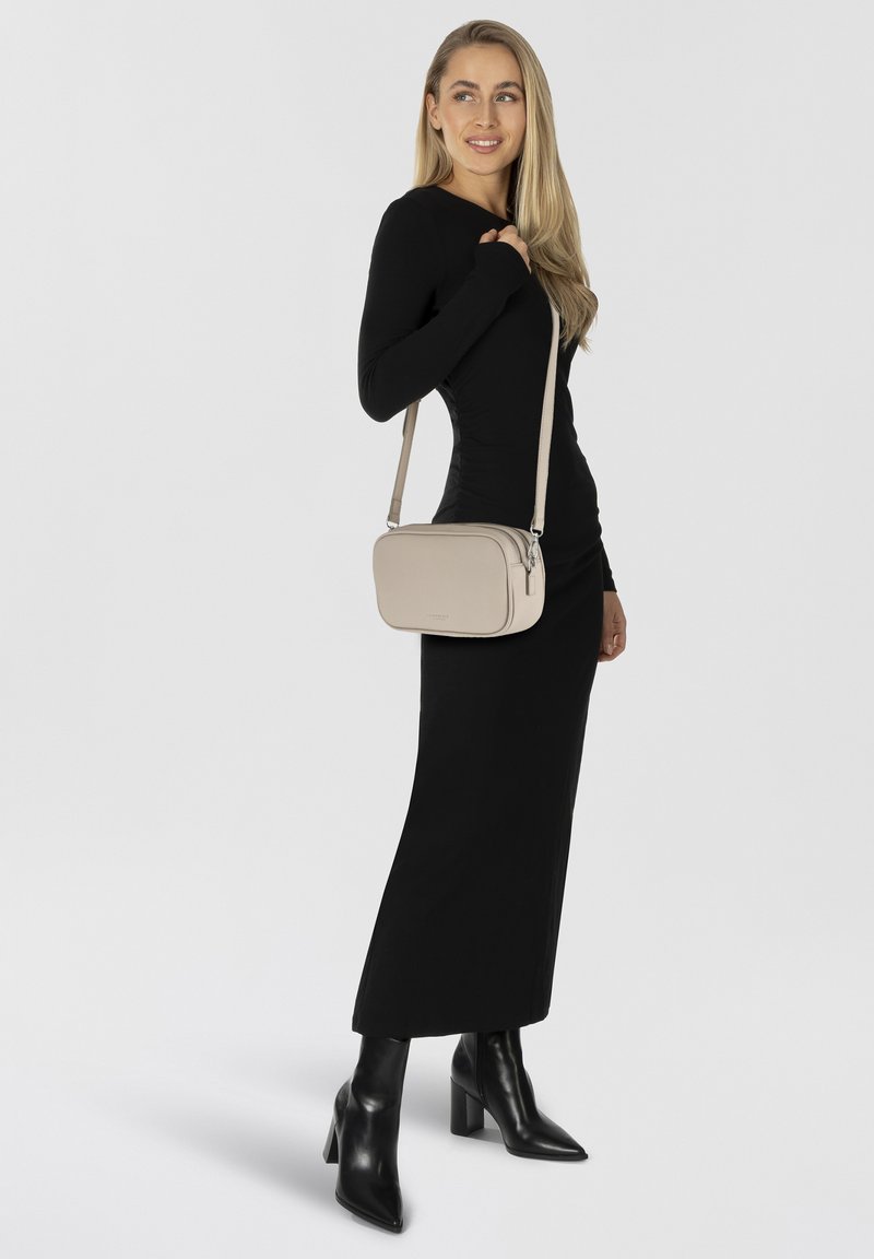 Beige Leder-Crossbody-Tasche mit glatter Textur, Reißverschluss und verstellbarem Trageriemen, getragen über einem schwarzen Langarmkleid und Ankle-Boots.