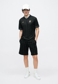 Νέος άνδρας που φοράει λευκό καπέλο, μαύρη κοντομάνικη φανέλα, μαύρα σορτς με κάθετο κείμενο, λευκές κάλτσες και λευκά αθλητικά παπούτσια, στέκεται χαλαρά.