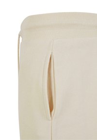Prim-plan cu pantaloni bej de trening, cu talie elastică cu striații, șnur și deschidere pentru buzunar lateral.
