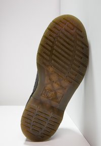 Semelle de chaussure en caoutchouc noir avec un grip texturé et des rainures horizontales, portant des détails de marque et de modèle embossés au centre.