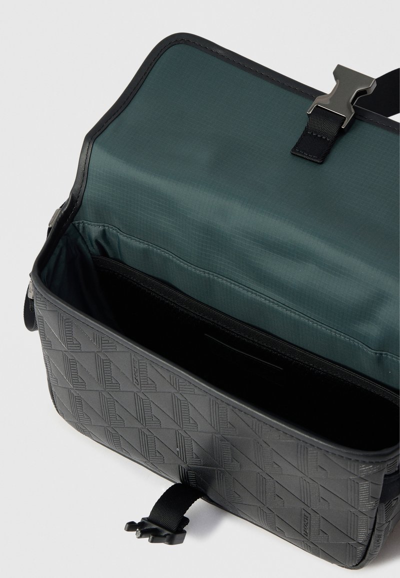 Sort tekstureret taske med geometrisk mønster, der har et grønt indre, lynlåslomme og lukning med sort plastspænde.