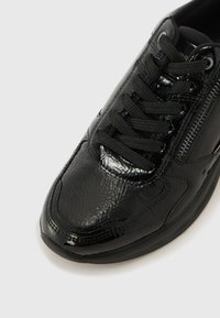 Baskets de sport noires avec une finition texturée et brillante, dotées de lacets noirs et d'une fermeture éclair sur le côté pour un enfilage facile. Bout rond et semelle flexible.
