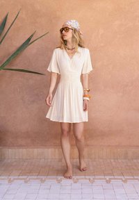 Robe enveloppante beige clair avec de courtes manches volantées, taille froncée et jupe fluide. Le mannequin est accessoirisé avec des bracelets colorés et des lunettes de soleil.