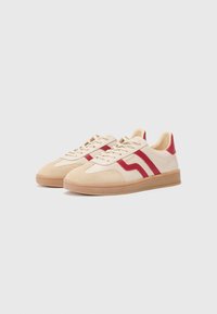 Baskets basses en cuir et daim beige avec des rayures rouges et une semelle en caoutchouc beige. Elles disposent de lacets et d'un col rembourré pour plus de confort.