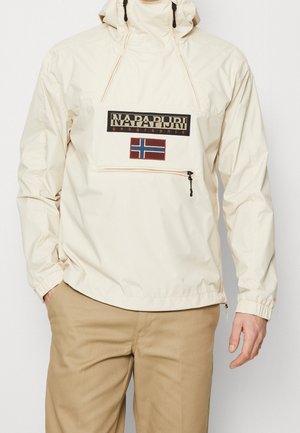 Mand iført en beige Napapijri pulloverjakke med forlommer med lynlås og norsk flagpatch, kombineret med lysebrune bukser.