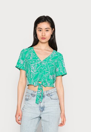 Top verde com mangas curtas e decote em V, apresentando um padrão floral em branco e rosa, atado à cintura. Combinado com jeans de tom azul claro.