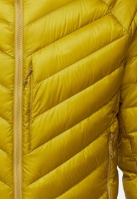 Veste doudoune jaune avec des sections diagonales en matelassage, texture lisse et fermeture avant zippée avec un tirette de contraste.