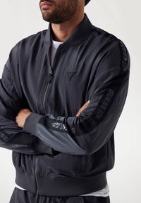 Black bomber jacket with a zip front, ribbed cuffs, and elastic hem. Features textured fabric and logo accent on the sleeves.