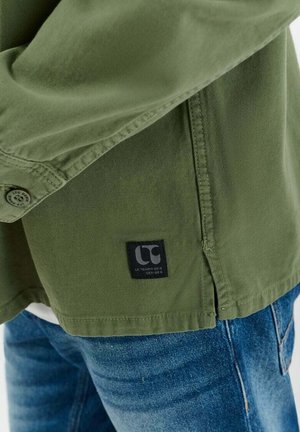 Close-up van de zoom van een groen overhemdd met knopen en een zwart merkembleem op blauwe denim jeans.