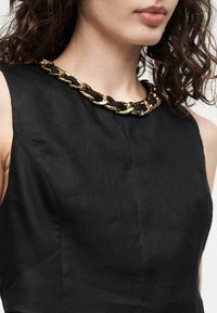 Γυναίκα με αμάνικο μαύρο φόρεμα, παχιά χρυσή και μαύρη αλυσίδα στο λαιμό, σγουρά σκούρα μαλλιά και φυσικά χείλη.