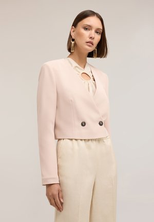 Frau trägt einen hellrosa kurzen Blazer mit zwei Knöpfen, eine cremefarbene Bluse mit Schlüssellochausschnitt, cremefarbene Hose und goldene Quastenohrringe.