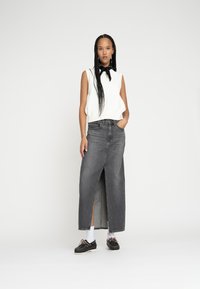 Gonna maxi in denim nero con spacco frontale, abbinata a un top senza maniche bianco e scarpe nere. La modella sta in piedi con una mano nella tasca.