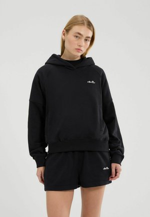 Młoda kobieta w czarnej bluzie z kapturem marki ellesse i dopasowanych spodenkach, stojąca na jednolitym szarym tle.