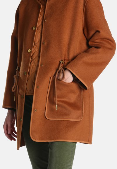 Manteau couleur rouille avec poches à cordon porté sur une veste matelassée orange brûlée et un pantalon vert, une main dans la poche.