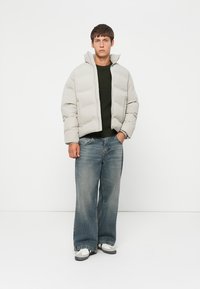 Lys grå pufferjakke med høj krave, lynlås foran og boxy fit. Båret over en mørkegrøn struktureret sweater og parret med løse denimjeans.