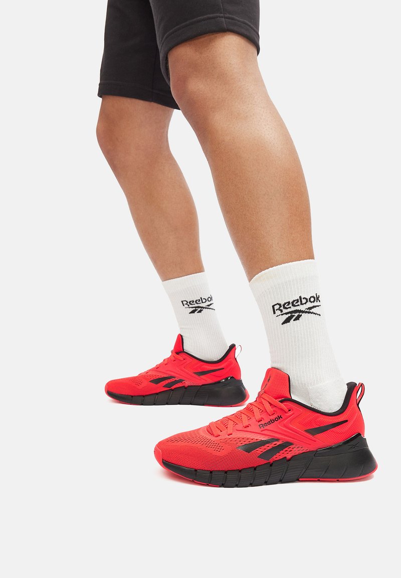 Scarpe sportive rosse con accenti neri, una parte superiore texturizzata e una suola spessa. Indossate con calzini bianchi con logo Reebok nero.