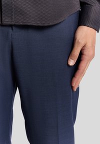 Kortärmad, texturerad mörk skjorta med ett geometriskt mönster, ihop med skräddarsydda mörkblå byxor. Handen vilar försiktigt på byxorna.