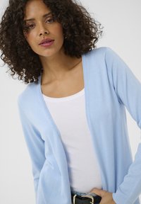 Hellblauer Strickcardigan mit offenem Vorderteil und langen Ärmeln, getragen über einem weißen Tanktop. Enthält einen schwarzen Gürtel mit einer goldenen Schnalle.