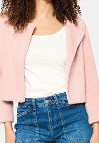 Jasnoróżowy, krótki kardigan o teksturowanym splocie, z otwartym przodem i rękawami do połowy przedramienia. Połączony z białym topem i niebieskimi, wysokim stanem jeansami.