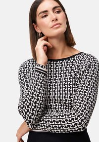 Zwart-wit gemusterde sweater met een geometrisch ontwerp, ronde hals en lange mouwen. Heeft een contrasterende gestreepte band bij de manchetten.