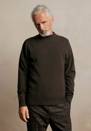 Brązowy dziany sweter z wysokim dekoltem, ściągaczami przy mankietach i u dołu. Noszony z wykrochmalonymi spodniami w kratę, z ciemnymi i jasnymi przetarciami.