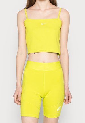 Kvinde iført en lys gul Nike cropped tanktop og matchende højttaljede cykelshorts, stående mod en ensfarvet baggrund.