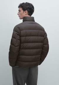 Giacca puffa marrone con trapuntatura orizzontale, colletto alto e una texture liscia, abbinata a pantaloni grigi e uno sfondo chiaro.