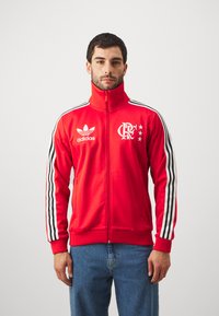 Red zip-up jacket with high collar, black and white striped sleeves, Adidas logo, and a team emblem featuring stars on the chest.